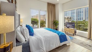 A modern hotel room with a large bed, a TV, and a view of the cityscape. - NEW 2BR Condo in  of Palm Beach! Walk 2 Beach! (Palm Beach)