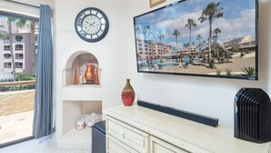 A hotel room with a large window, a wall-mounted clock, a framed picture of a beach, a small shelf with decorative items, and a wooden dresser.