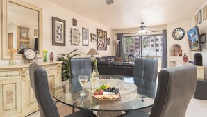 A dining area with a glass table surrounded by chairs, a bottle of wine, and a fruit platter.