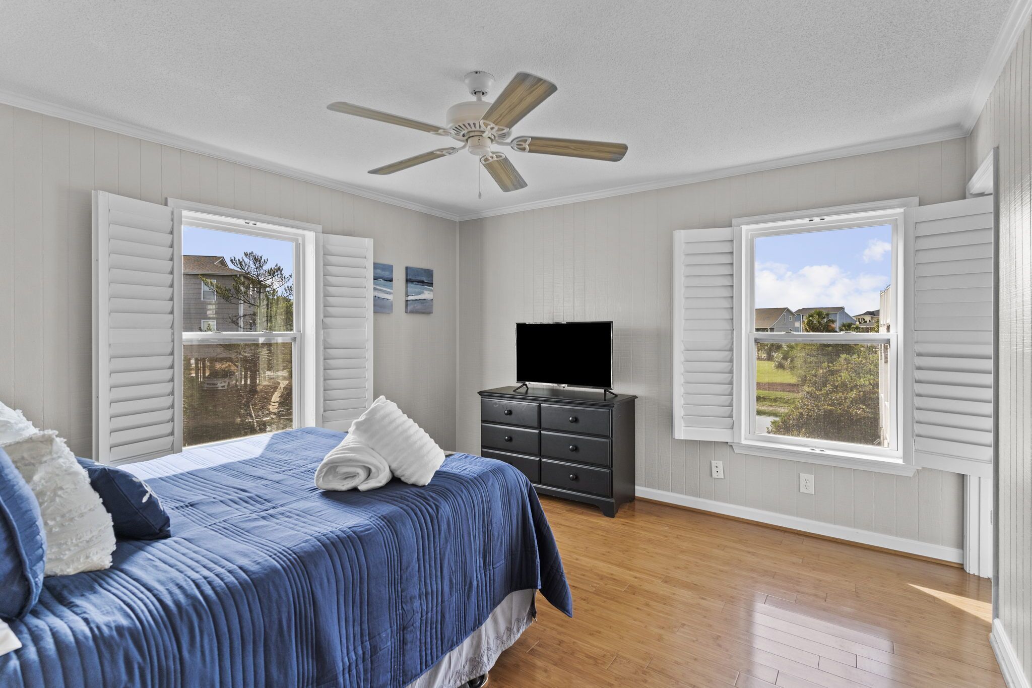 A bedroom with a bed, a TV, a ceiling fan, and windows with shutters.
