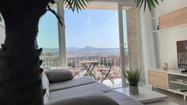 A modern living room with a sofa, a small table, and a TV. There is a balcony with a view of the city and mountains.