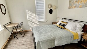 Un dormitorio con cama, ventilador de techo, ventana con persianas, una mesita pequeña y una silla.