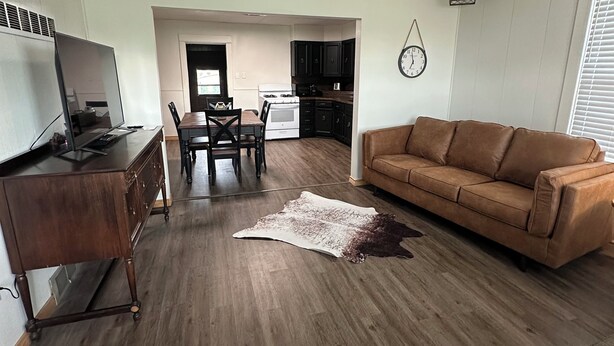 Una sala de estar con un sofá de cuero marrón, una mesa de centro de madera y un televisor sobre un mueble de madera. Hay un comedor con mesa y sillas, y una cocina al fondo.