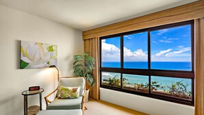 Una habitación con un ventanal grande con vista al océano, una silla con cojín, una mesita con un reloj y una planta en maceta.