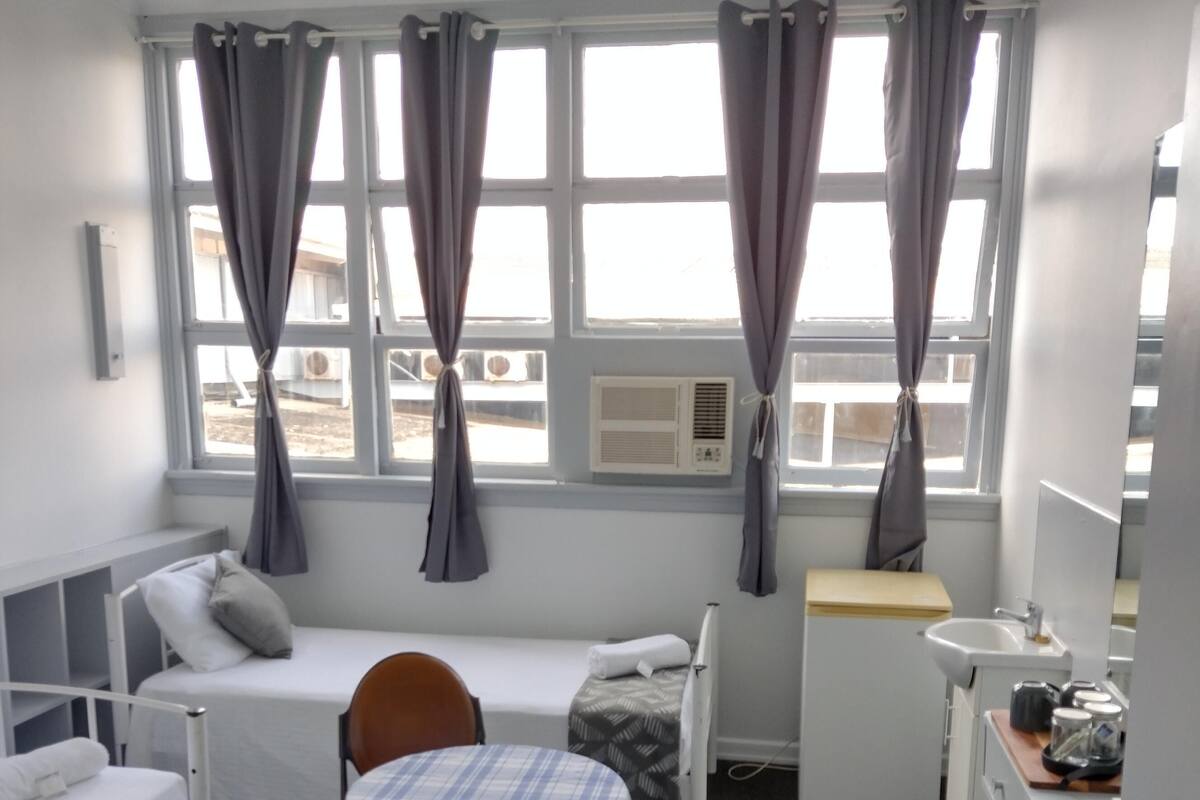 A compact hotel room with a single bed, a small table with a chair, a desk with a chair, and a window with curtains.
