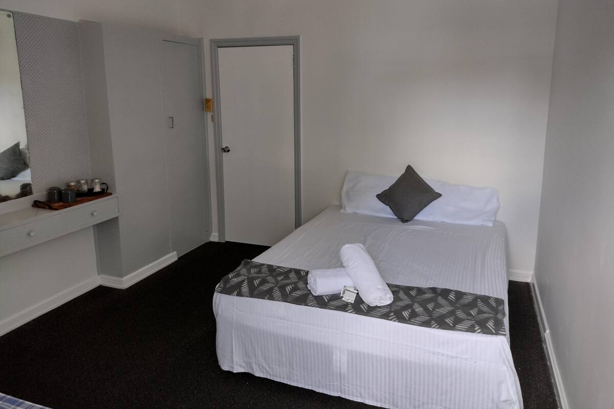 A hotel room with a bed, a bedside table with a mirror, a door, and a small shelf with jars.