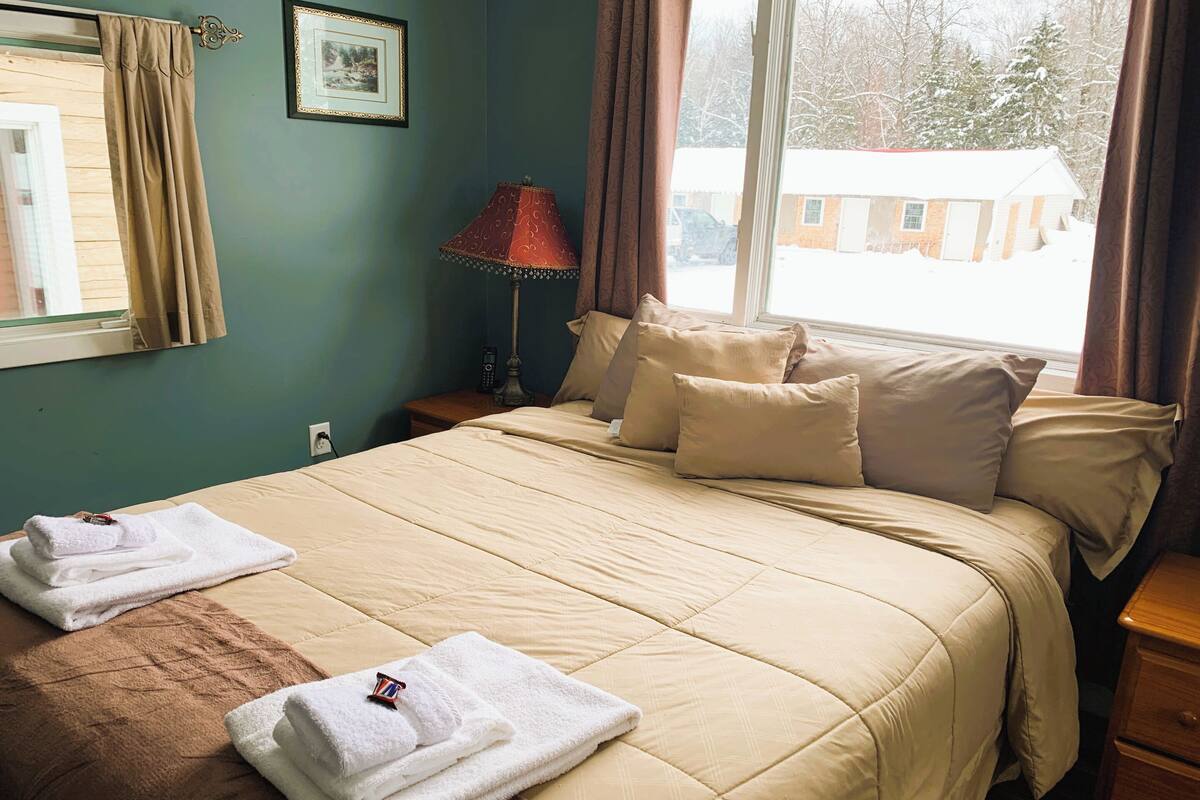 Ett hotellrum med en säng, kuddar, en lampa, ett fönster med gardiner och handdukar.
