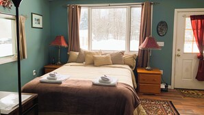 Une chambre à coucher comprenant un lit, deux tables de chevet, une lampe, une fenêtre avec des rideaux et une porte avec un paillasson.