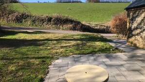A paved patio with a round table, a stone wall, and a grassy field with sheep in the background.