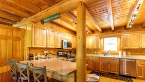Interior de una cabaña de madera con zona de cocina, que incluye armarios de madera, una isla central con sillas, un microondas y un lavavajillas de acero inoxidable.