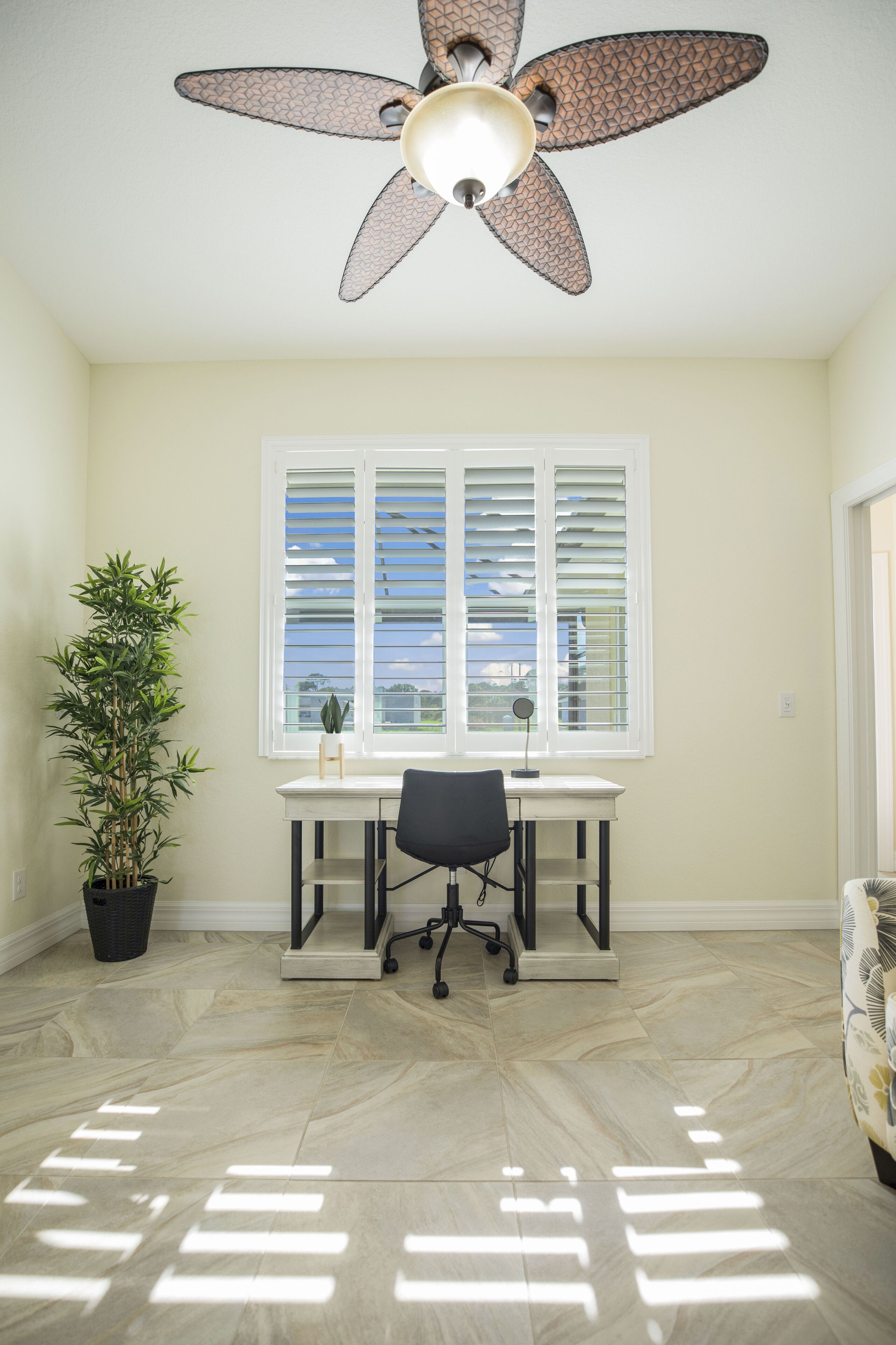 Ein Zimmer mit Deckenventilator, Schreibtisch mit Stuhl, Fenster mit Jalousien und einem Blumentopf.