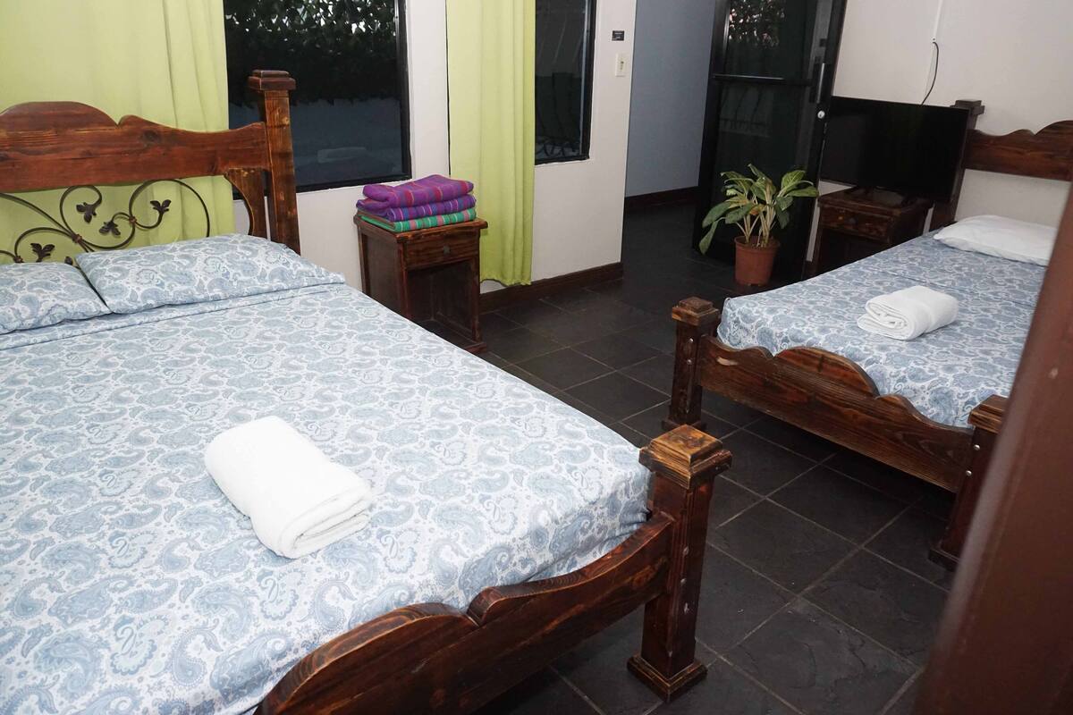 Habitación de hotel con dos camas de madera, cada una con una colcha estampada y una toalla enrollada. Hay una mesita de noche de madera con una planta y una manta doblada.