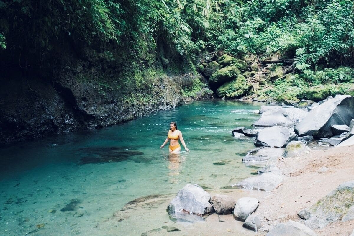 A woman in a yellow swimsuit standing in a clear river surrounded by rocks and greenery.