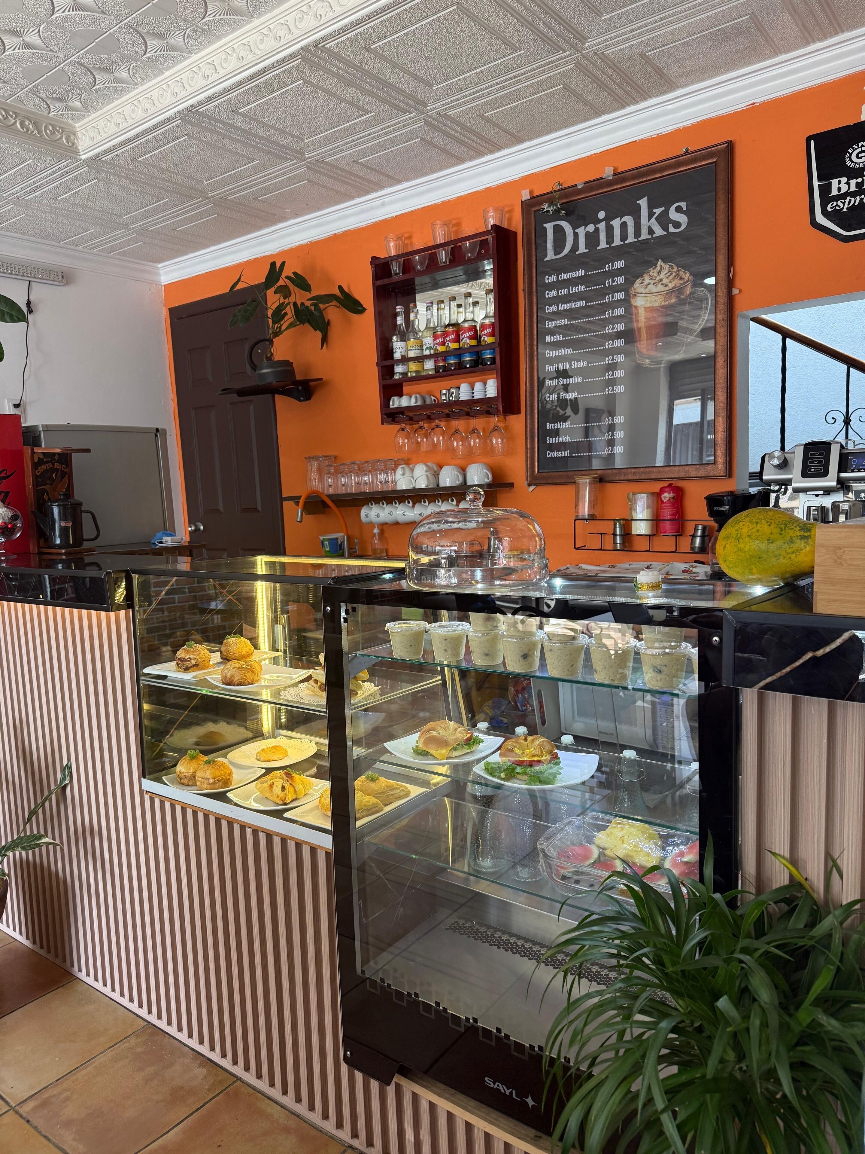 Una cafetería con una vitrina llena de pasteles y sándwiches, un menú de bebidas en la pared y una máquina de café al fondo.