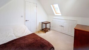 Un dormitorio con techo inclinado, tragaluz, cama con manta marrón, pequeña mesa y armario.