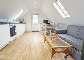 Un loft moderno con cocina, comedor y sala de estar. Cuenta con piso de madera, armarios blancos, un sofá gris y una mesa de centro de vidrio.