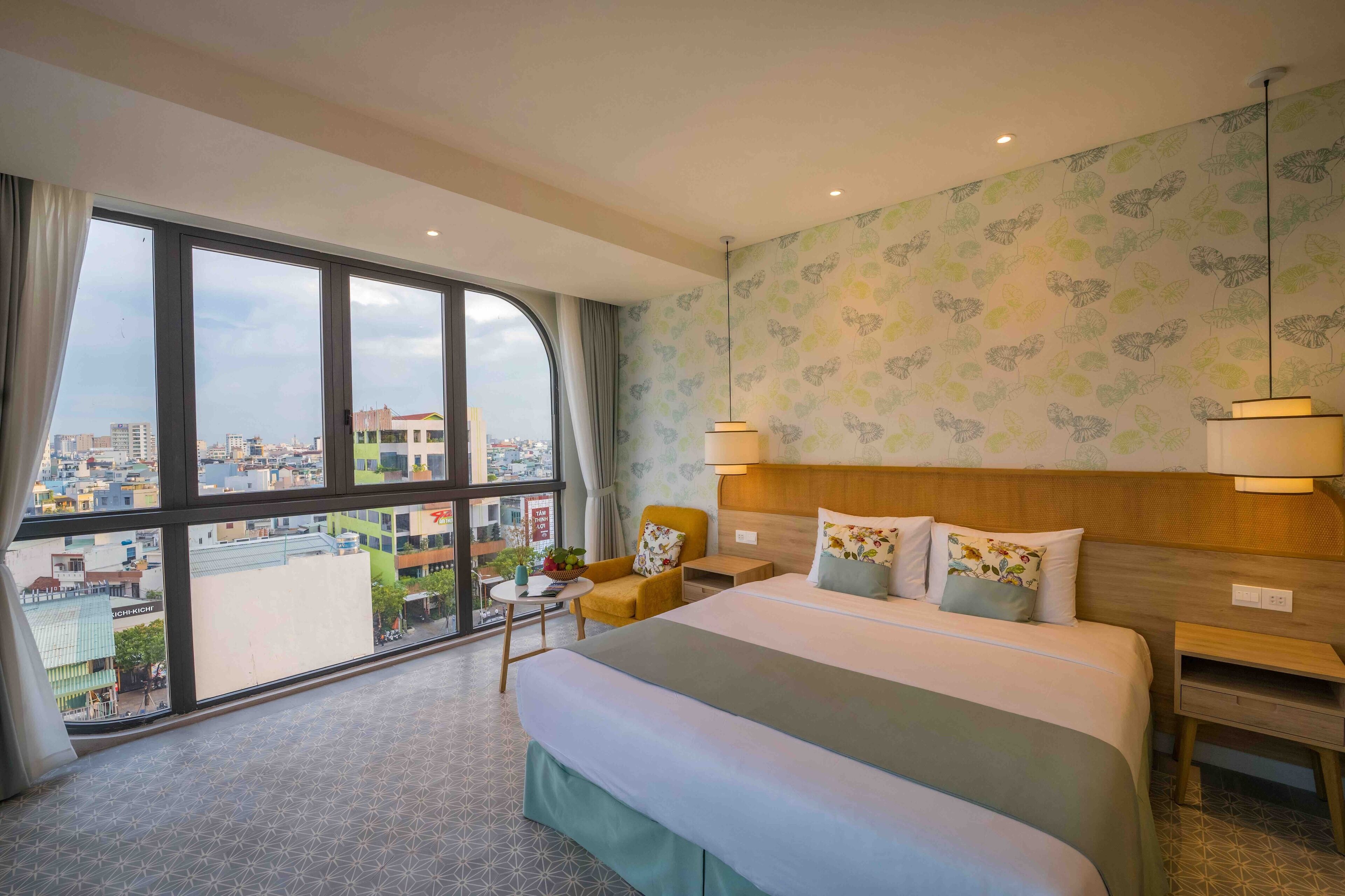 A hotel room with a large bed, two bedside tables, a nightstand, a chair, a small table with a plant, and a view of the city through large windows.