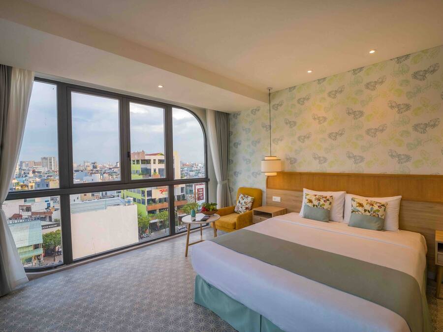 A hotel room with a large bed, two bedside tables, a nightstand, a chair, a small table with a plant, and a view of the city through large windows.