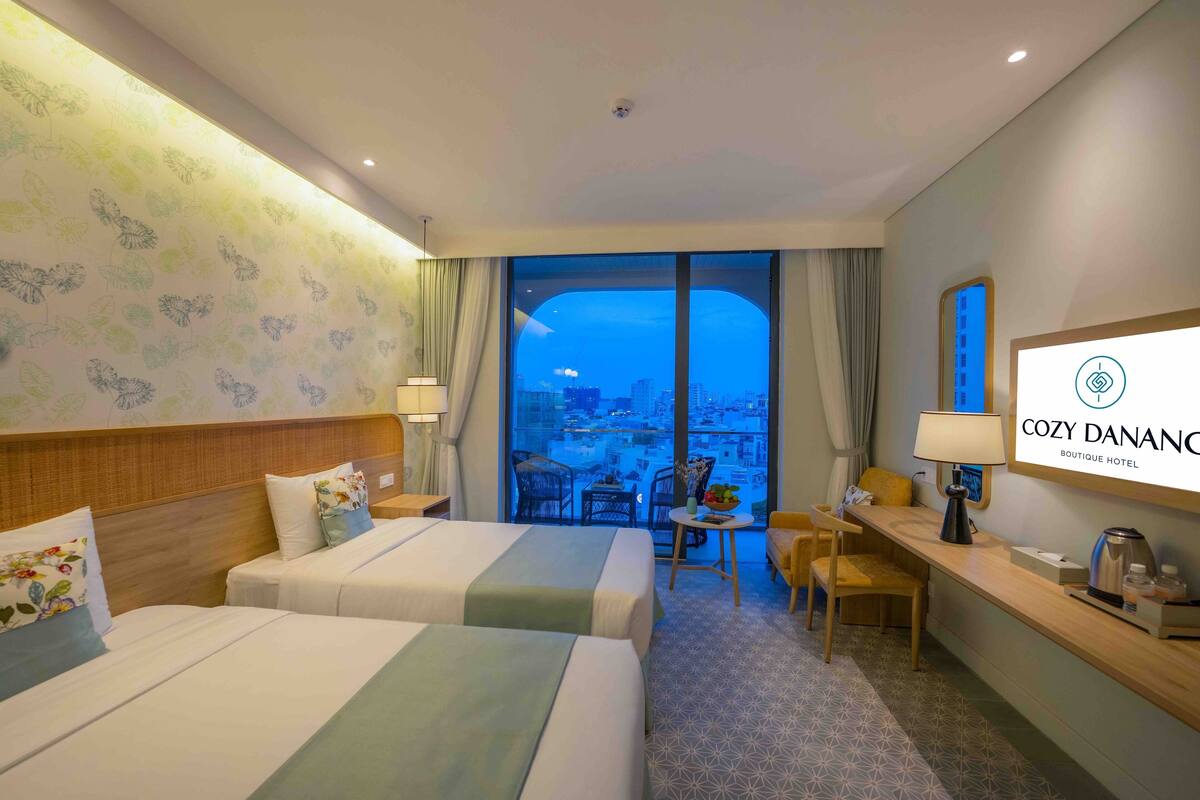 A hotel room with two beds, a desk, a chair, a small table with a lamp, and a large window offering a city view.