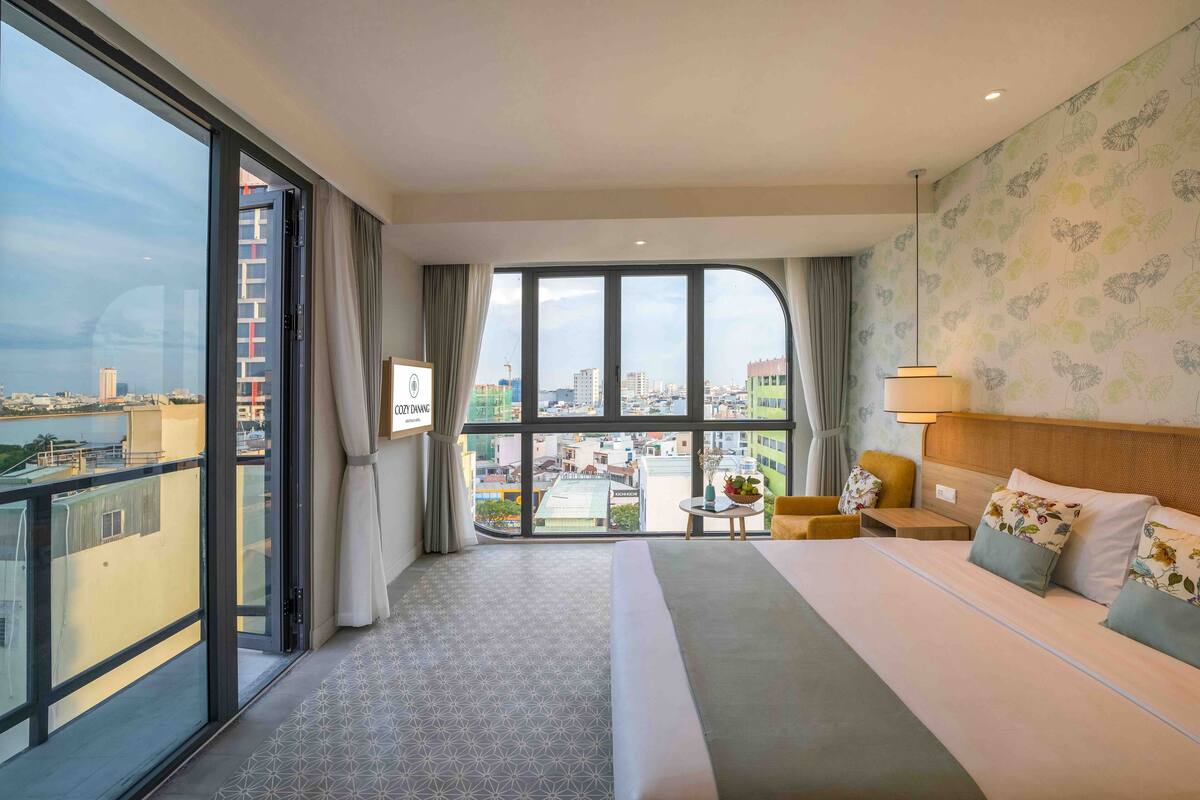 A hotel room with a large bed, a sofa, a small table, and a view of the city through large windows.