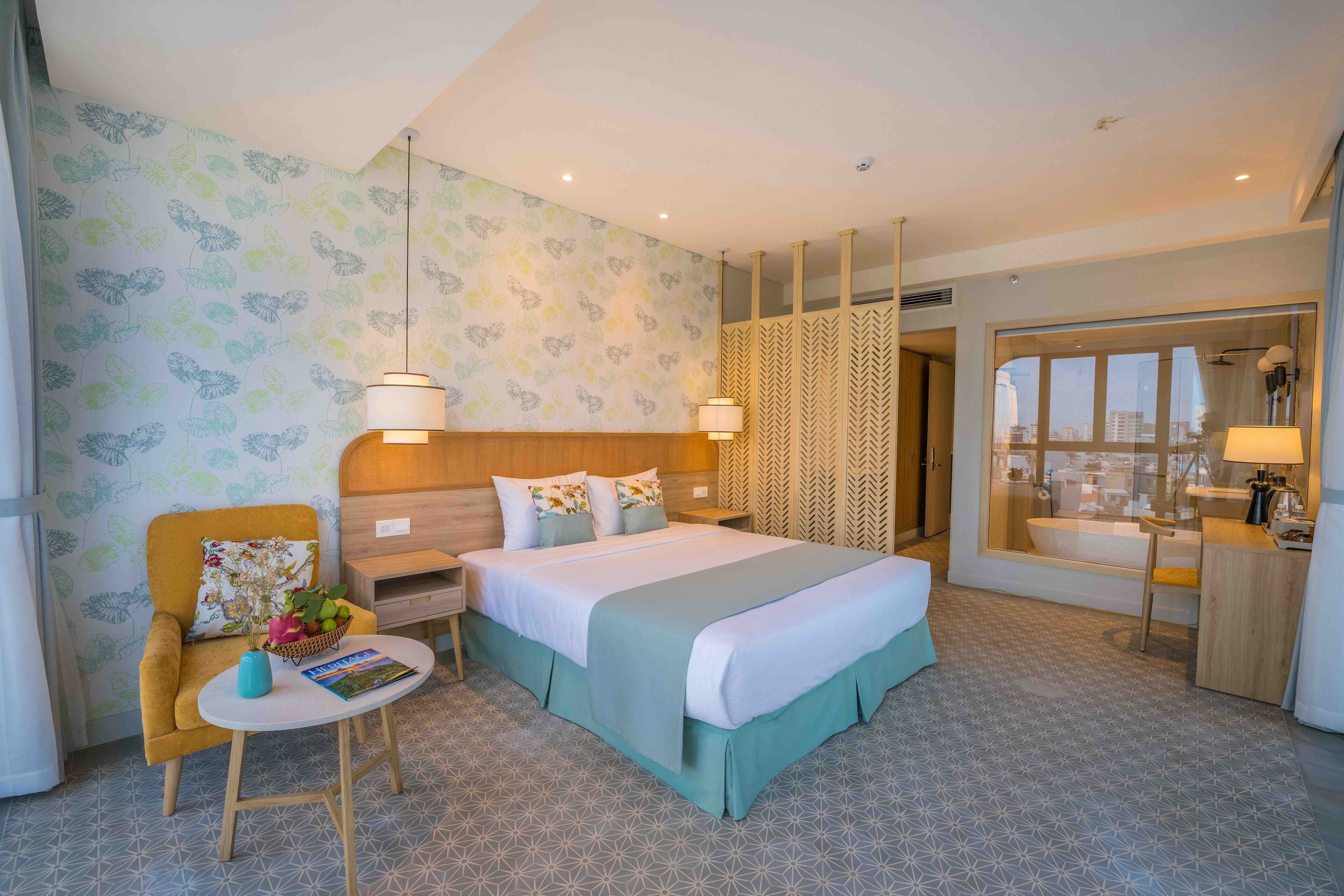 A hotel room with a bed, a chair, a small table, a nightstand, a mirror, and a bathroom visible through a glass door.