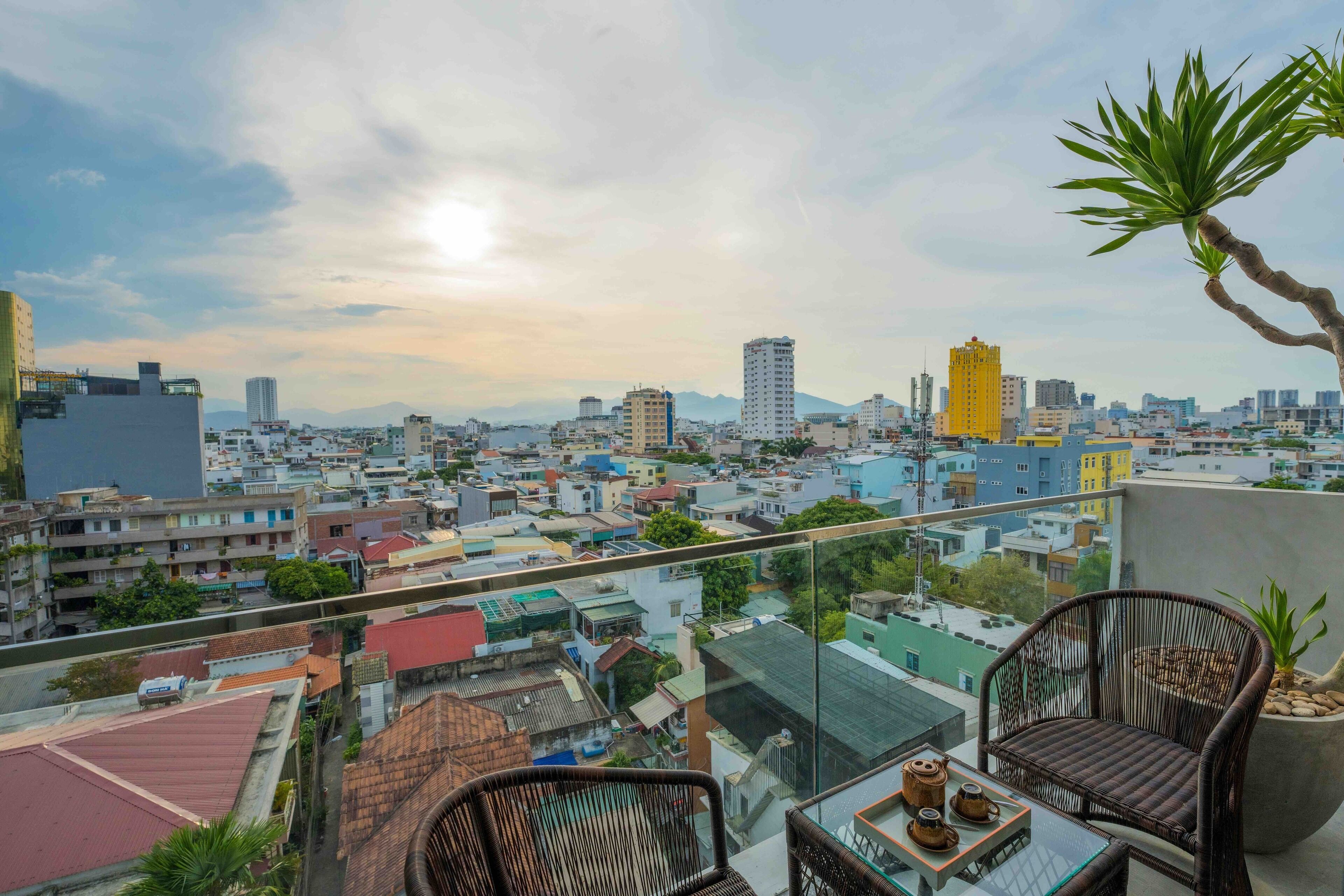A balcony with wicker chairs and a table, overlooking a cityscape with various buildings and a clear sky.