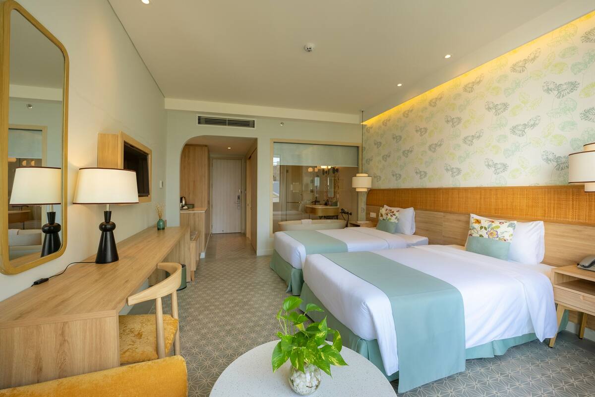 A hotel room with two beds, a desk with a chair, a round table with a plant, and a large mirror.
