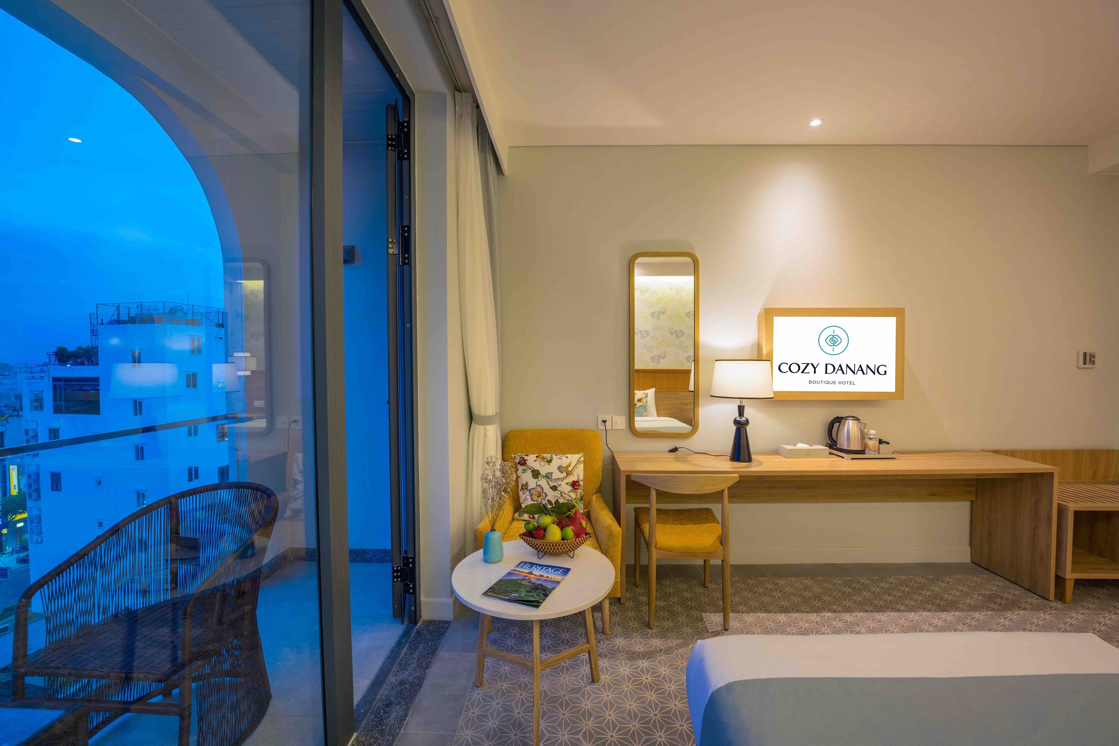 A hotel room with a balcony, a desk, a chair, a bed, and a small table with a magazine.