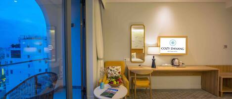 A hotel room with a balcony, a desk, a chair, a bed, and a small table with a magazine.
