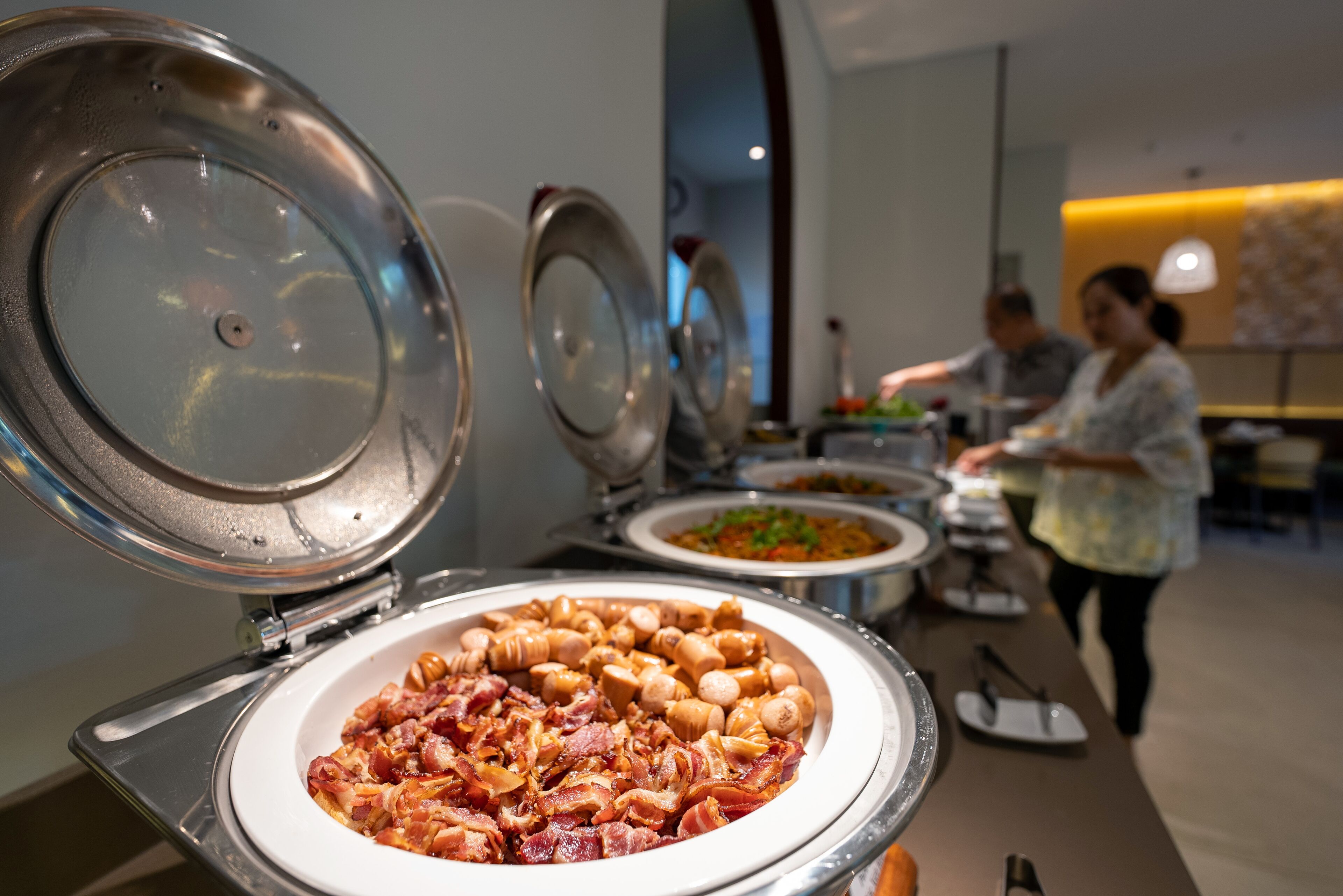 A buffet with various dishes, including a large plate of bacon and mushrooms, and a person serving food in the background.