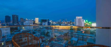A high-rise balcony with wicker furniture and a table, overlooking a cityscape with buildings and a green-lit structure in the distance.
