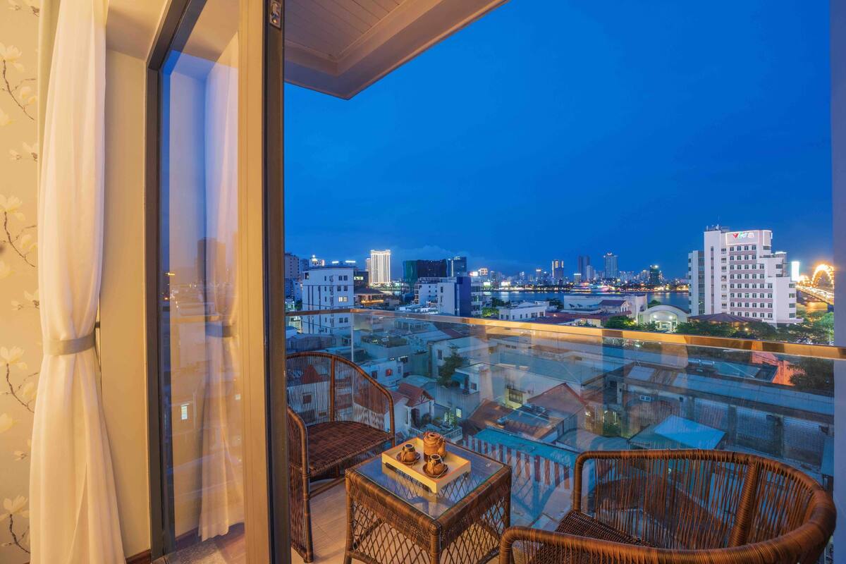 A balcony with wicker furniture and a table, overlooking a cityscape at dusk.