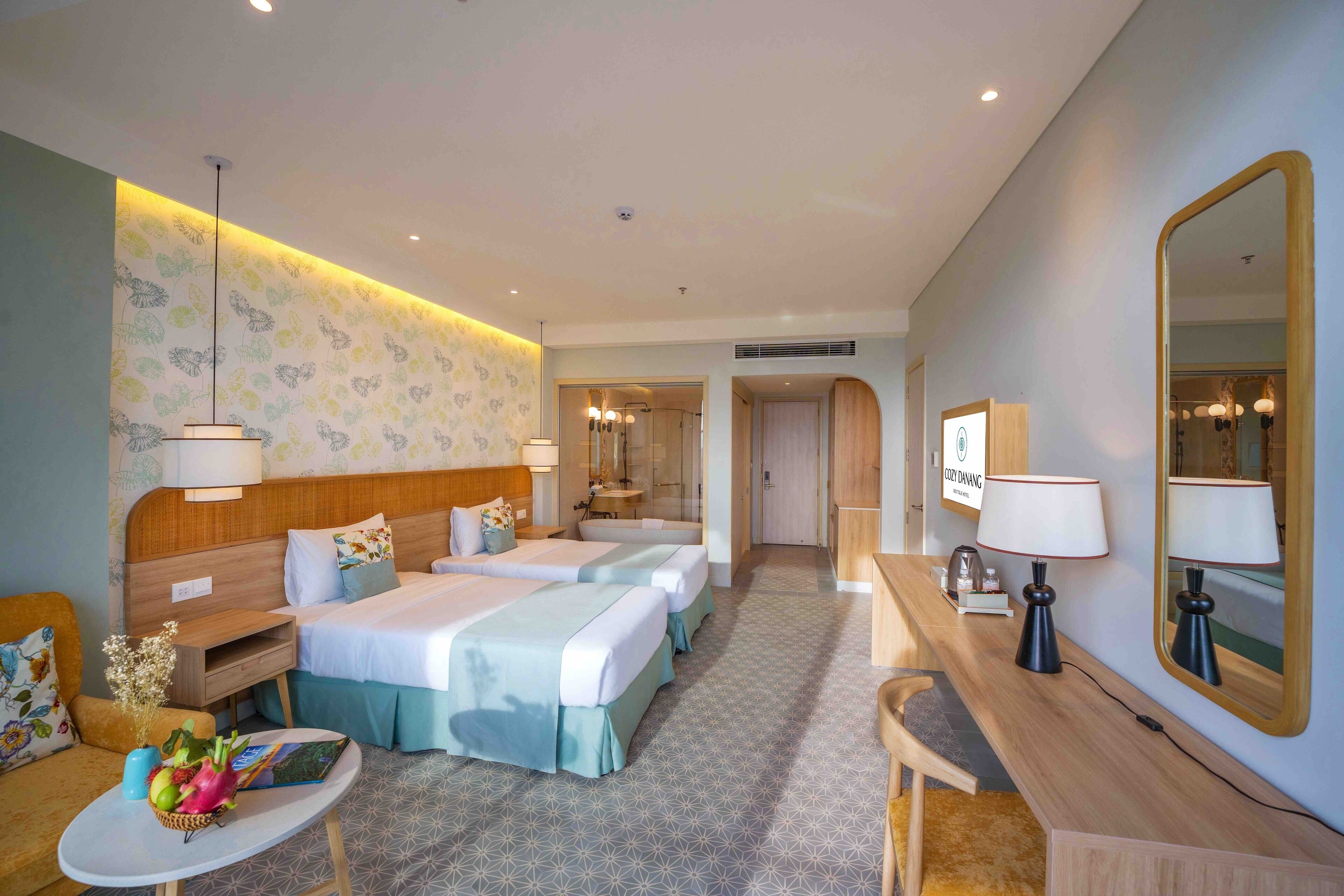 A hotel room with two beds, a desk, a chair, a small table with a vase of flowers, and a mirror.