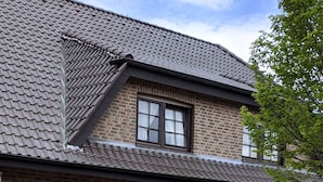 Ein zweigeschossiges Backsteinhaus mit einem Ziegeldach, einer Veranda mit zwei Holzstühlen und einem Garten mit verschiedenen Pflanzen und Blumen.