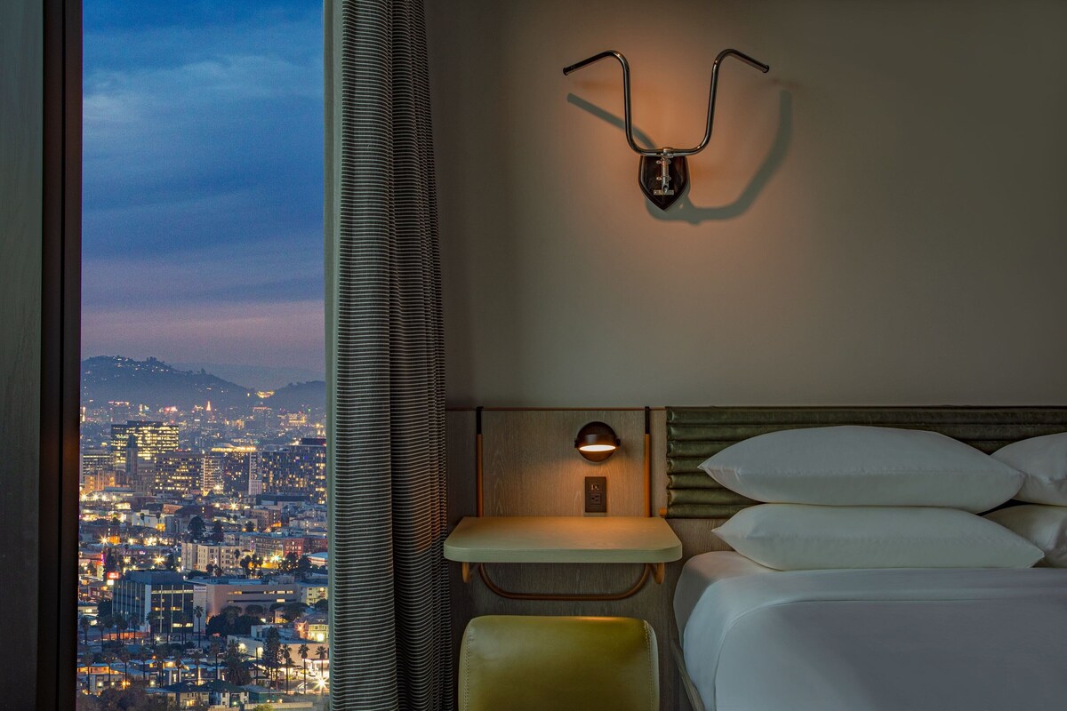 Habitación de hotel con vista a la ciudad, cama con ropa blanca, mesita de noche con lámpara y cabecera con lámpara.