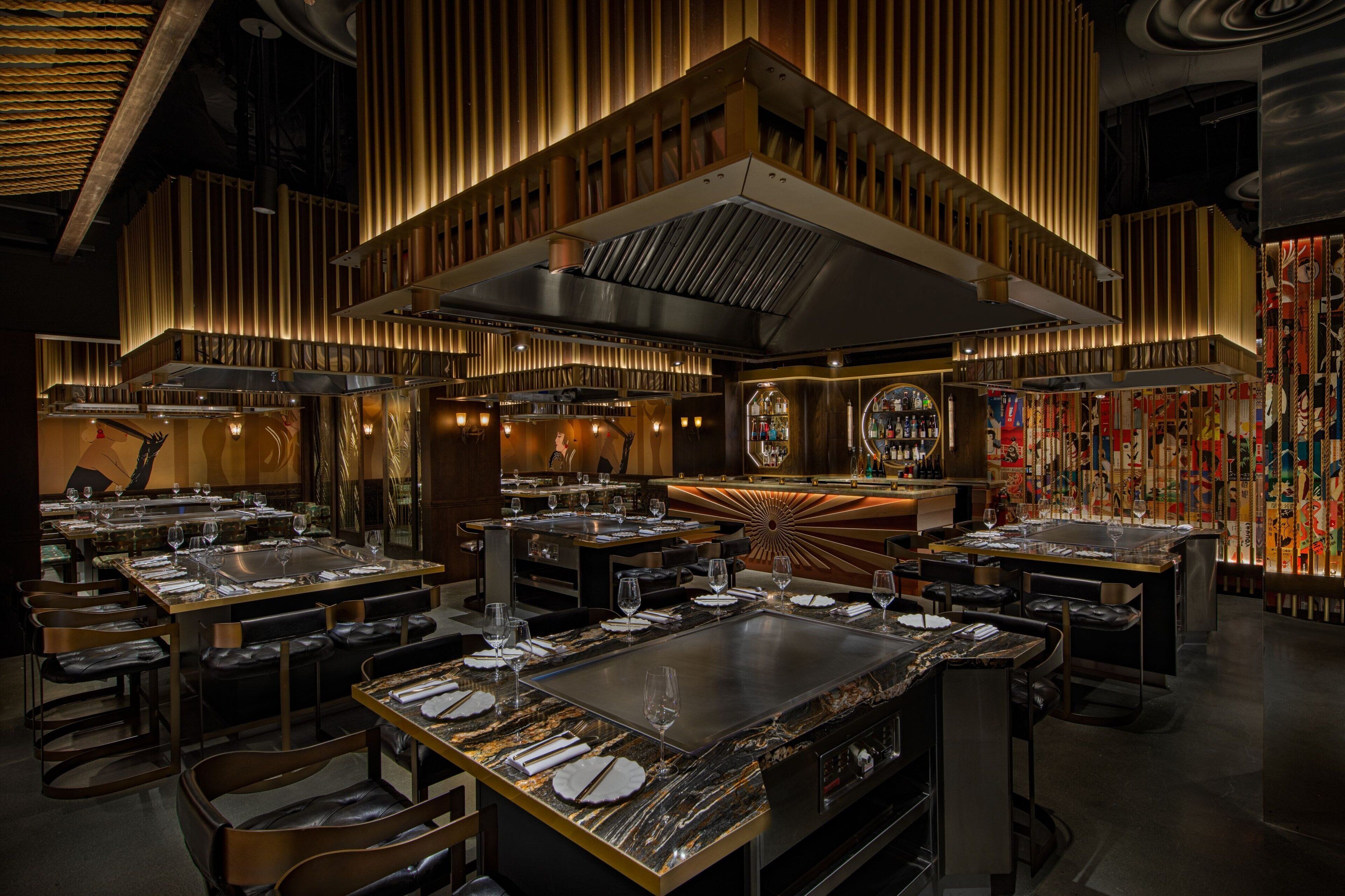 A modern restaurant interior with multiple dining tables, a central bar area, and a decorative wall with hanging elements.