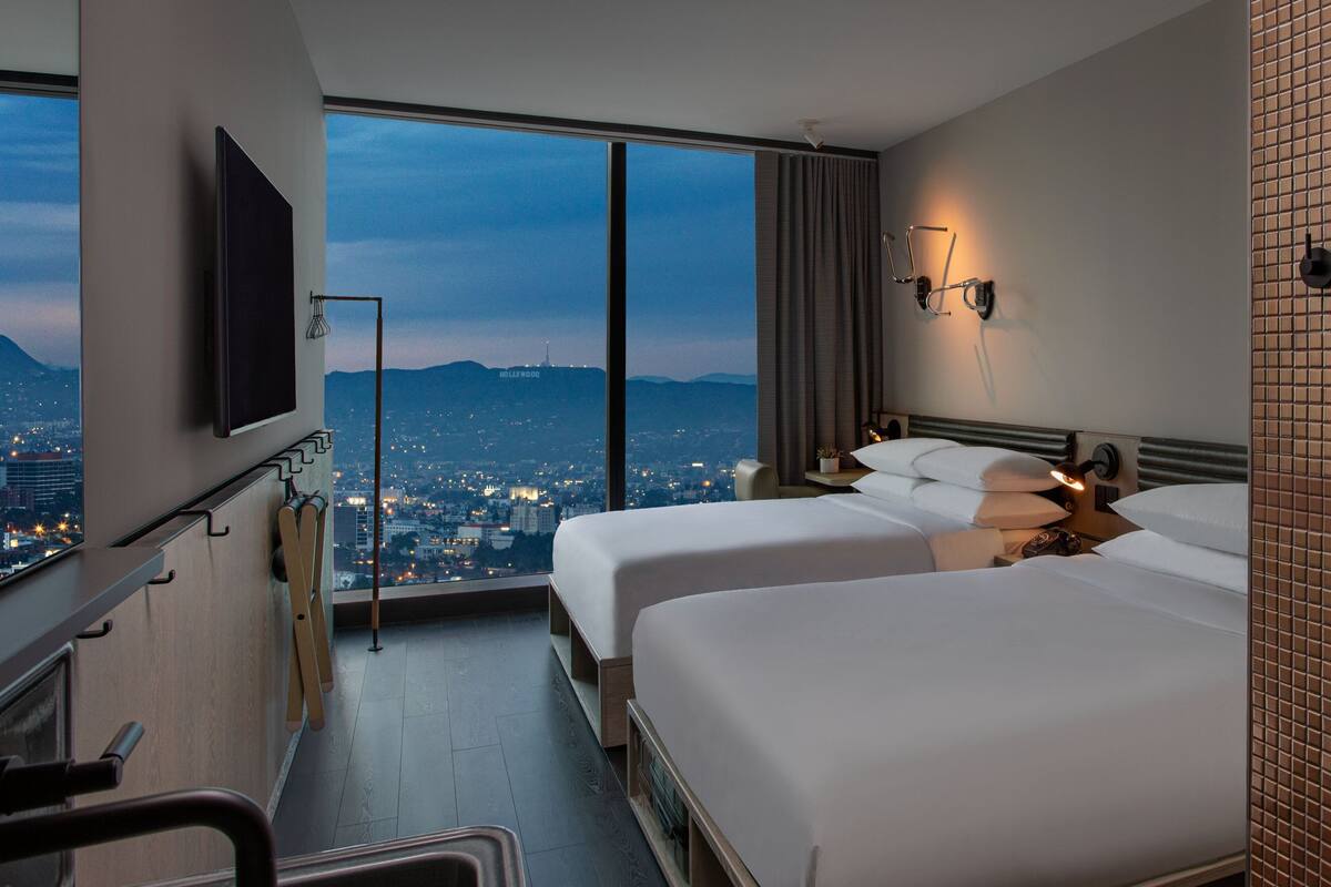 Ein Hotelzimmer mit zwei Betten, einem Fernseher, Blick auf die Stadt und einer Schiebetür zum Balkon.