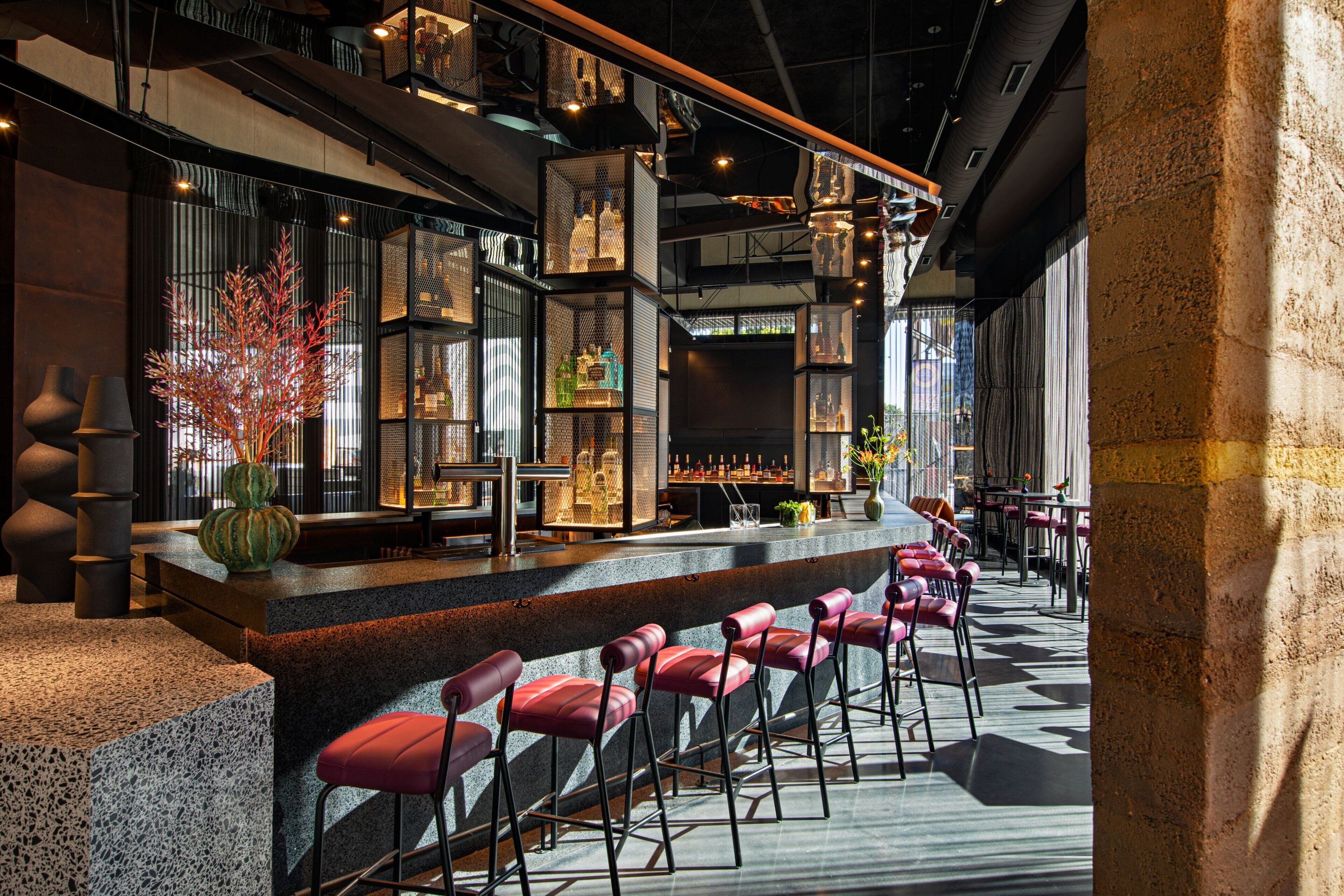 A modern bar area with a long counter, red bar stools, and shelves stocked with bottles and glasses.