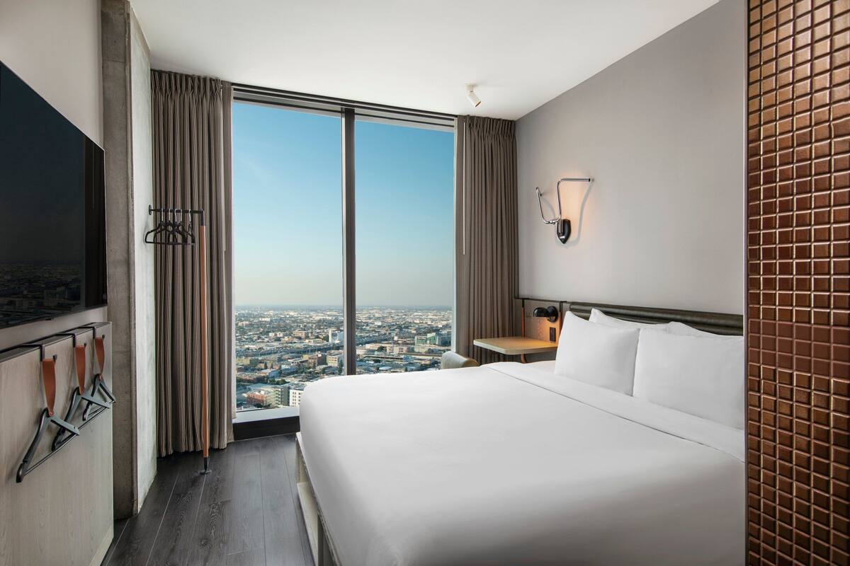Ein Hotelzimmer mit einem großen Bett, einem an der Wand befestigten Fernseher, Blick auf die Stadt und einem mit Vorhängen versehenen Fenster.