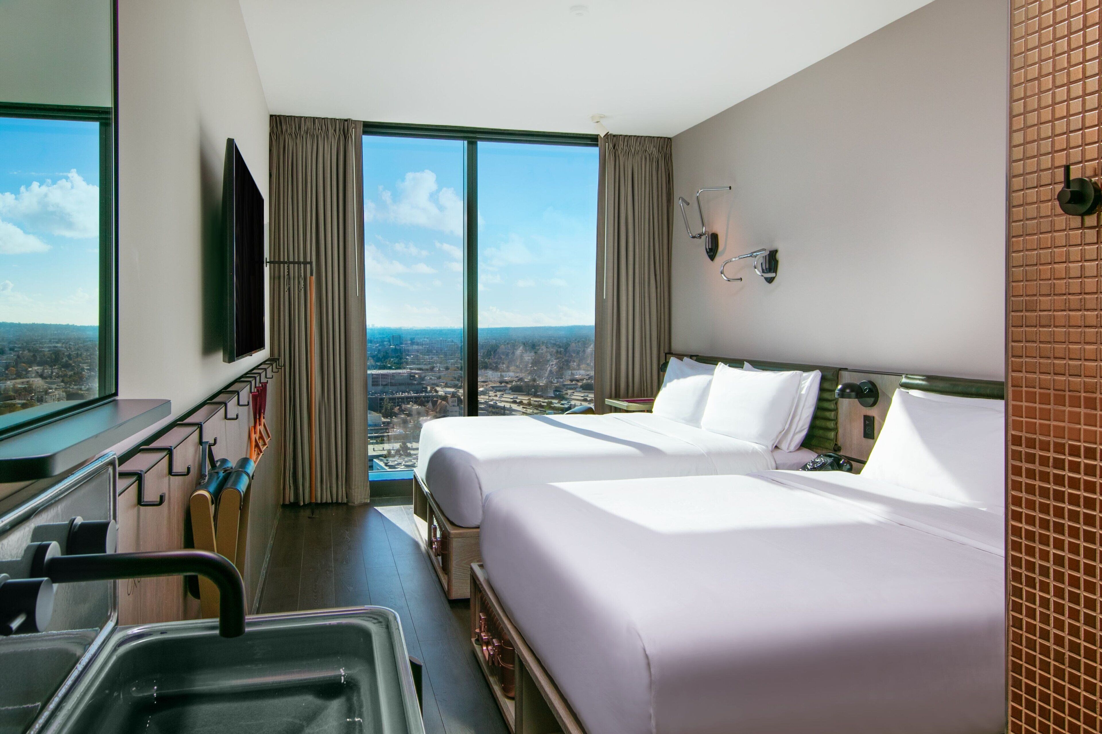 Quarto de hotel com duas camas, uma janela grande com vista para a cidade, televisão e banheiro com espelho e porta-toalhas.