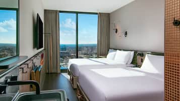 Quarto de hotel com duas camas, uma janela grande com vista para a cidade, televisão e banheiro com espelho e porta-toalhas.