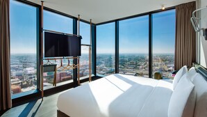 Habitación de hotel con una cama grande, un televisor sobre un soporte y vistas a la ciudad a través de amplias ventanas.