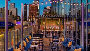 Un espacio al aire libre con mesas y sillas, una zona de bar con un muro decorado, y un horizonte urbano de fondo.