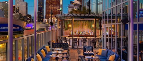 Un espacio al aire libre con mesas y sillas, una zona de bar con un muro decorado, y un horizonte urbano de fondo.