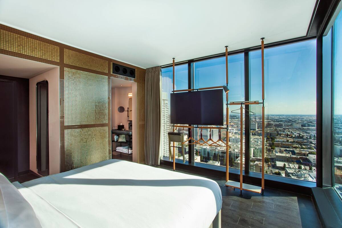 Ein Hotelzimmer mit einem großen Bett, einem an einer Metallkonstruktion befestigten Fernseher und einem Blick auf die Stadt durch große Fenster.
