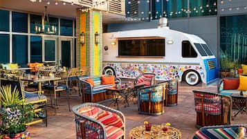 Un patio al aire libre con mobiliario de mimbre, un camión de comida y un edificio con ventanas amplias al fondo.