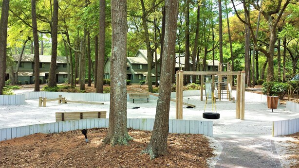 Un parque infantil con columpios y un tobogán, rodeado de árboles y un edificio al fondo.