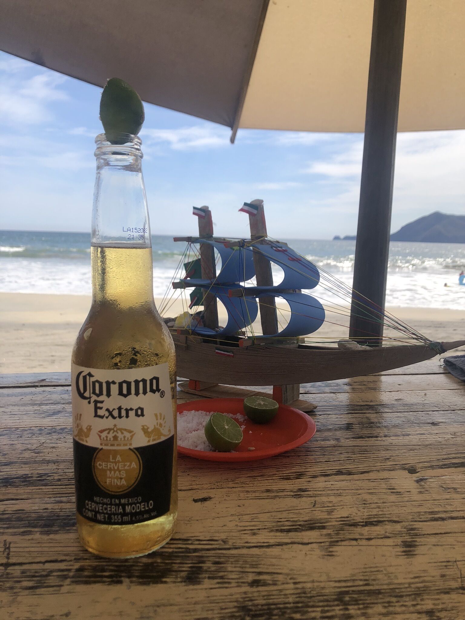 A bottle of Corona Extra beer on a wooden table with a hammock and a red plate with lime wedges.