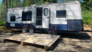 A travel trailer with a covered deck area, two folding chairs, and a small table. - Trailer on Spacious lakefront Campground (Lone Butte)