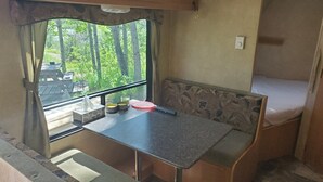 A compact RV interior with a small table, bench seating, a window with curtains, and a bed. - Trailer on Spacious lakefront Campground (Lone Butte)